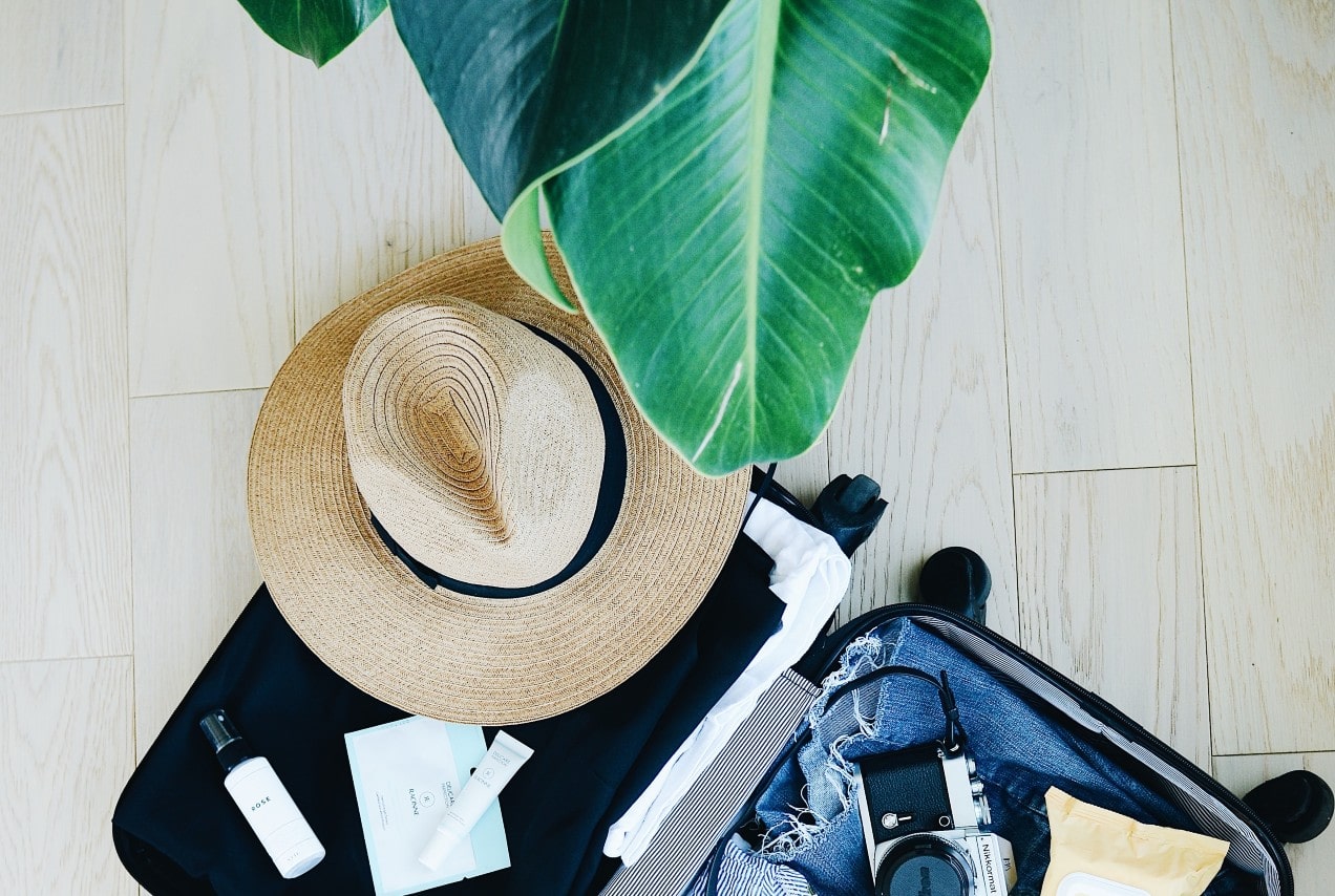 An open suitcase full of clothes, creams, a camera, and a hat, on top of the bright wooden floor.