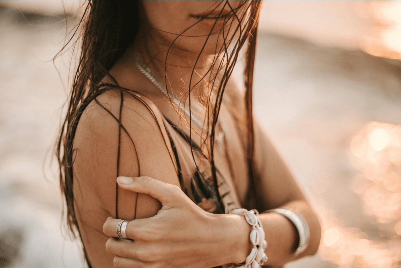 A woman with wet hair on the beach, showcasing fine jewelry pieces, including a shell bracelet and stackable rings.