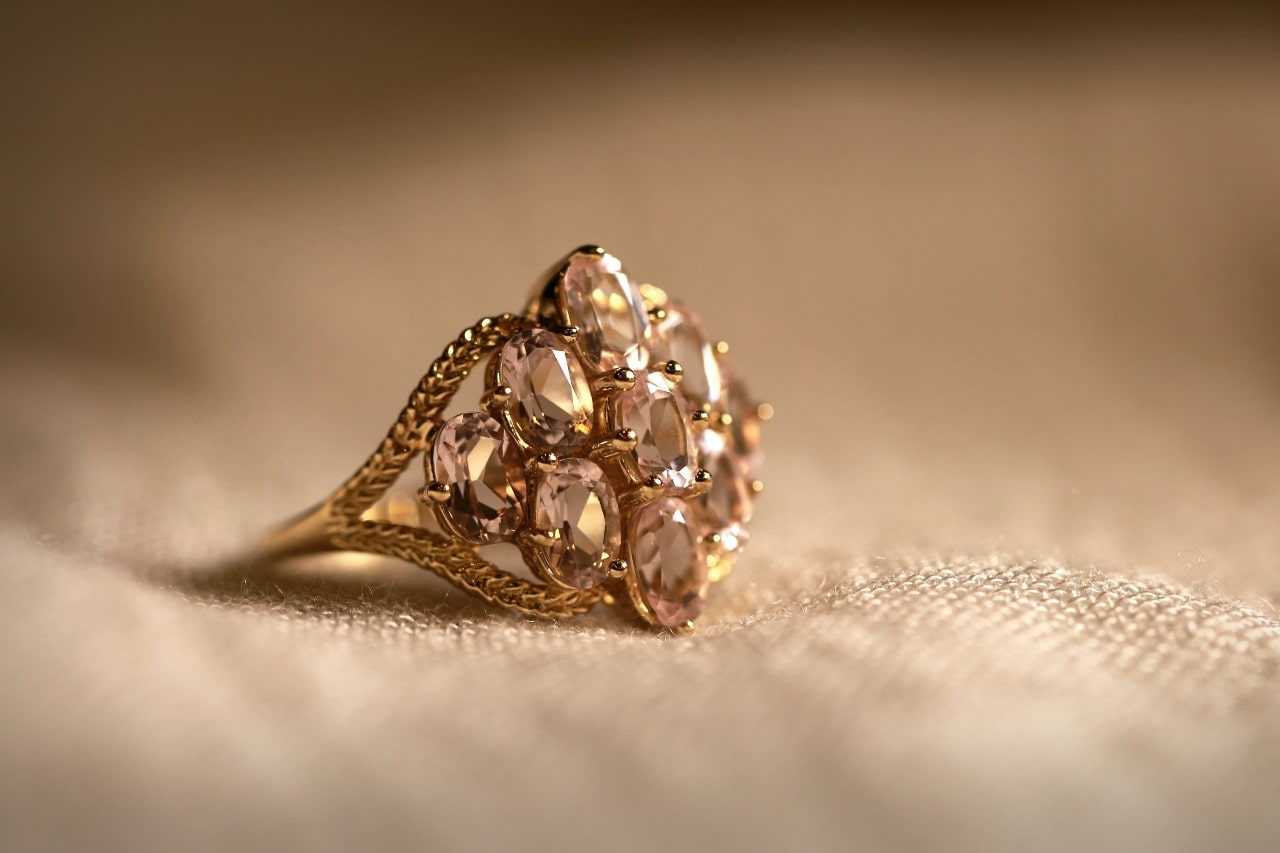 A vintage-style gold ring with multiple oval-cut light pink gemstones is displayed on a beige fabric surface.
