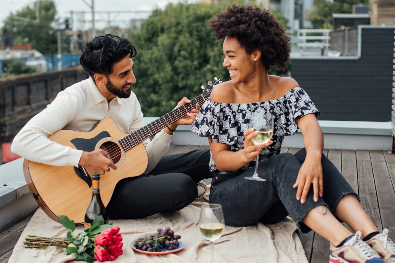 Couple sits in a balcony: man plays a guitar while woman holds a glass of wine.