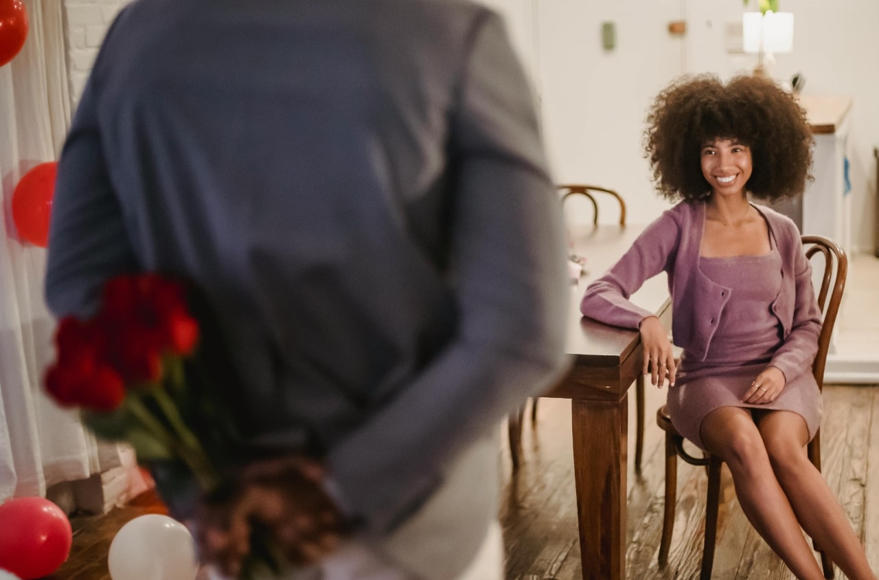 A woman in a lilac dress sits on a table while men in a gray shirt facing her, while hiding a bouquet of red roses behind his back.