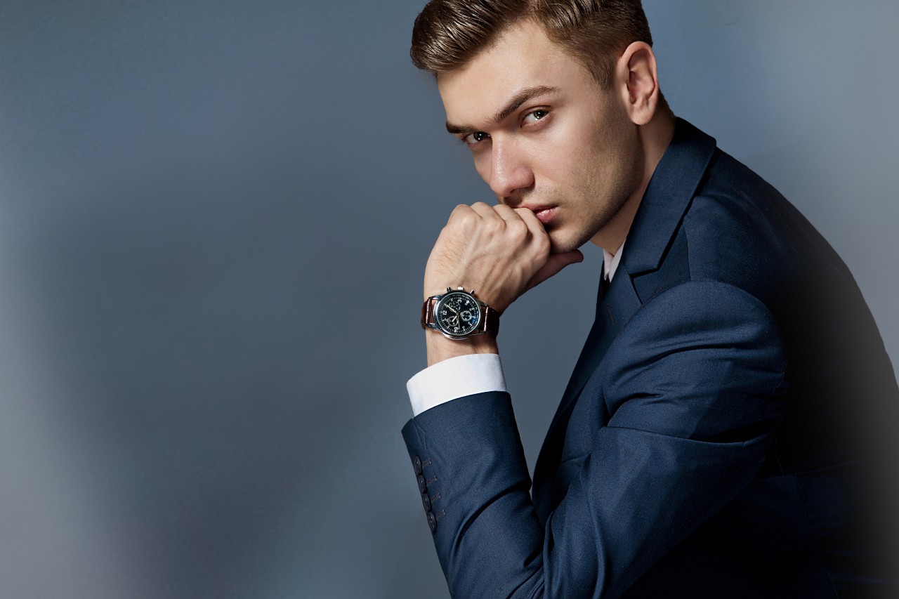 A young man in a navy suit gazes confidently at the camera, resting his chin on his hand, clad in a stylish luxury watch.