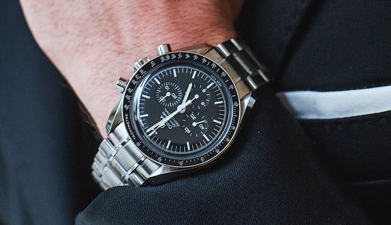 A sleek silver chronograph watch with a black dial and intricate markings is worn on a wrist in formal attire.