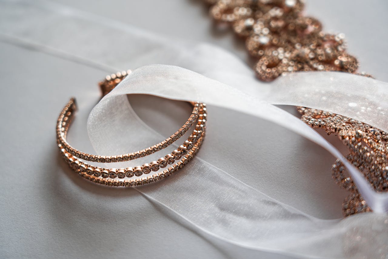 Rose gold crystal-studded bangles rest on soft white fabric with a sheer ribbon and matching sparkling jewelry in the background.