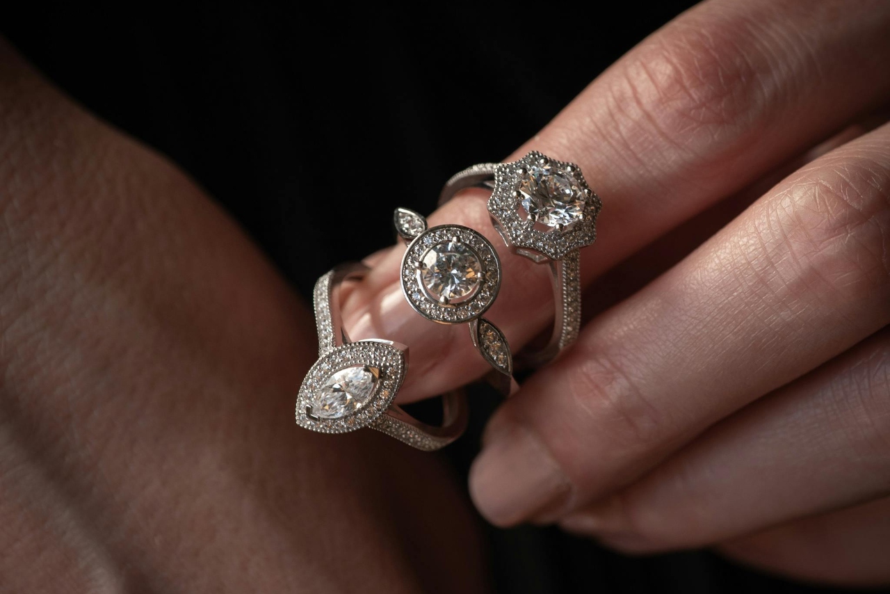 Three elegant diamond rings with intricate settings, held by fingers against a soft, dark background
