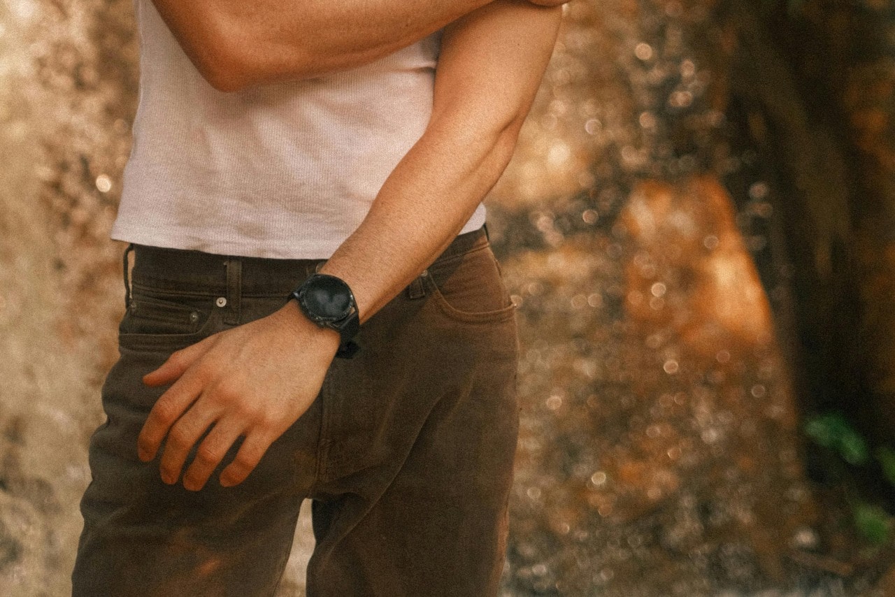 Close-up of a man wearing a black smartwatch, a white tank top, and brown jeans.