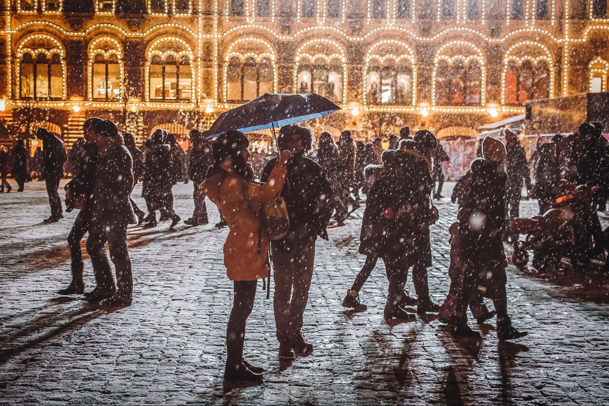 A crowd of people walks across a snowy, cobblestone square at night, illuminated by warm golden lights from an ornate historic building, with one couple standing under an umbrella in the front.