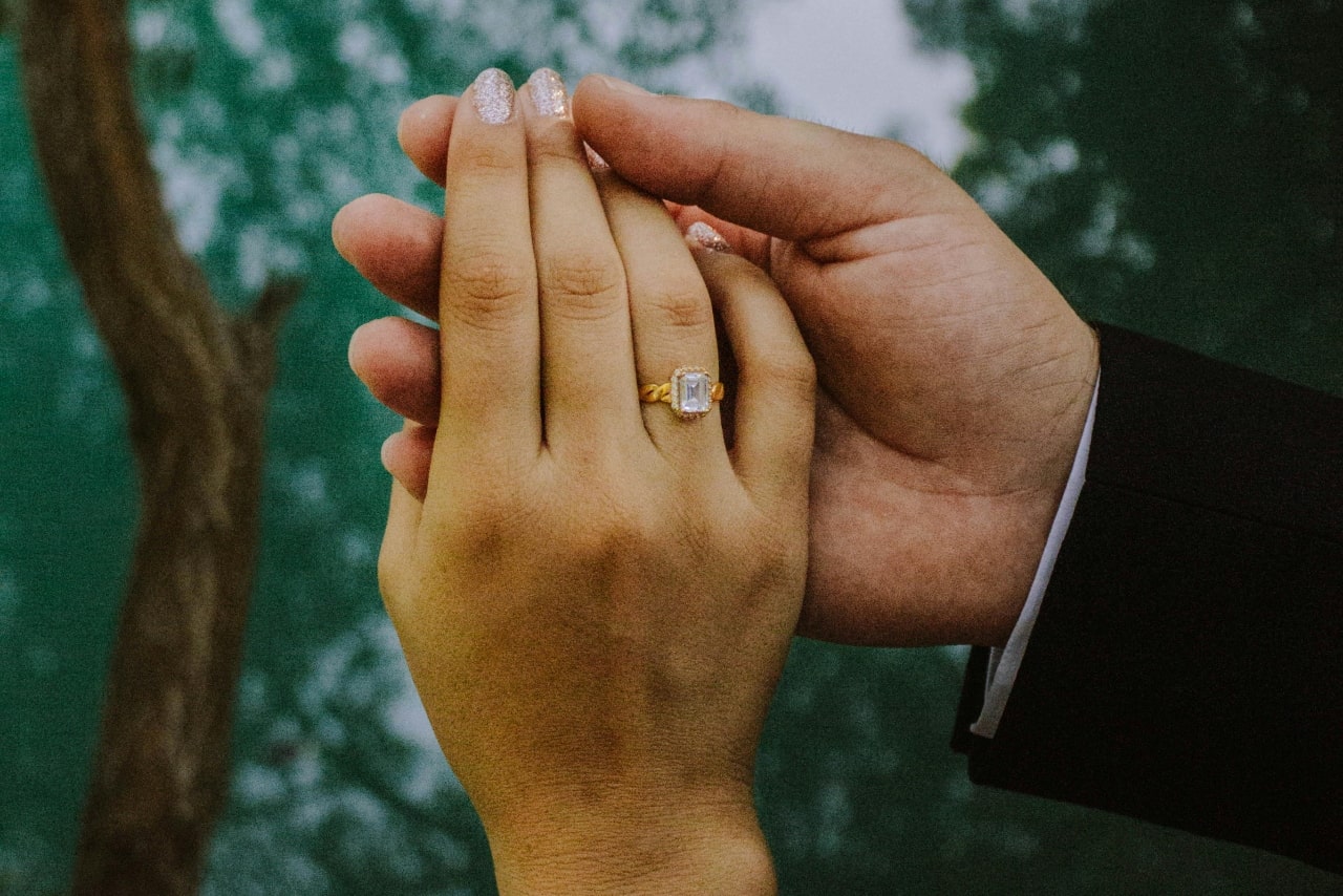 A couple holding hands, the woman’s hand adorned with a yellow gold emerald cut engagement ring and sparkly nails.