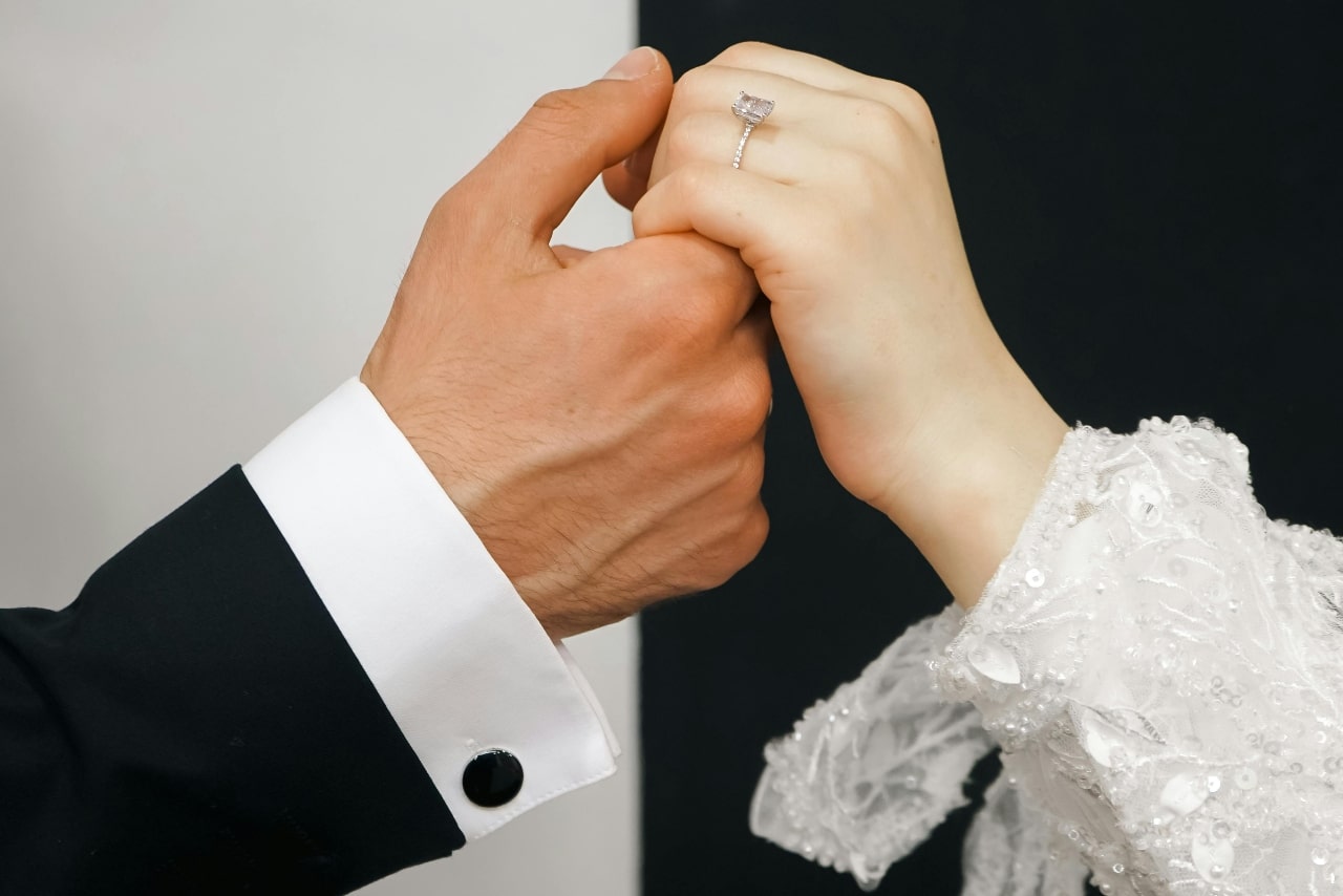 A man and woman in their wedding attire holding hands, the woman’s hand adorned with a side stone engagement ring.