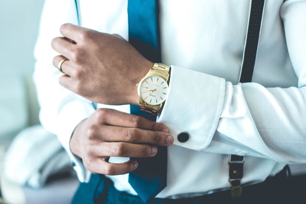 A man in a white shirt and blue tie adjusts a gold watch on his wrist.