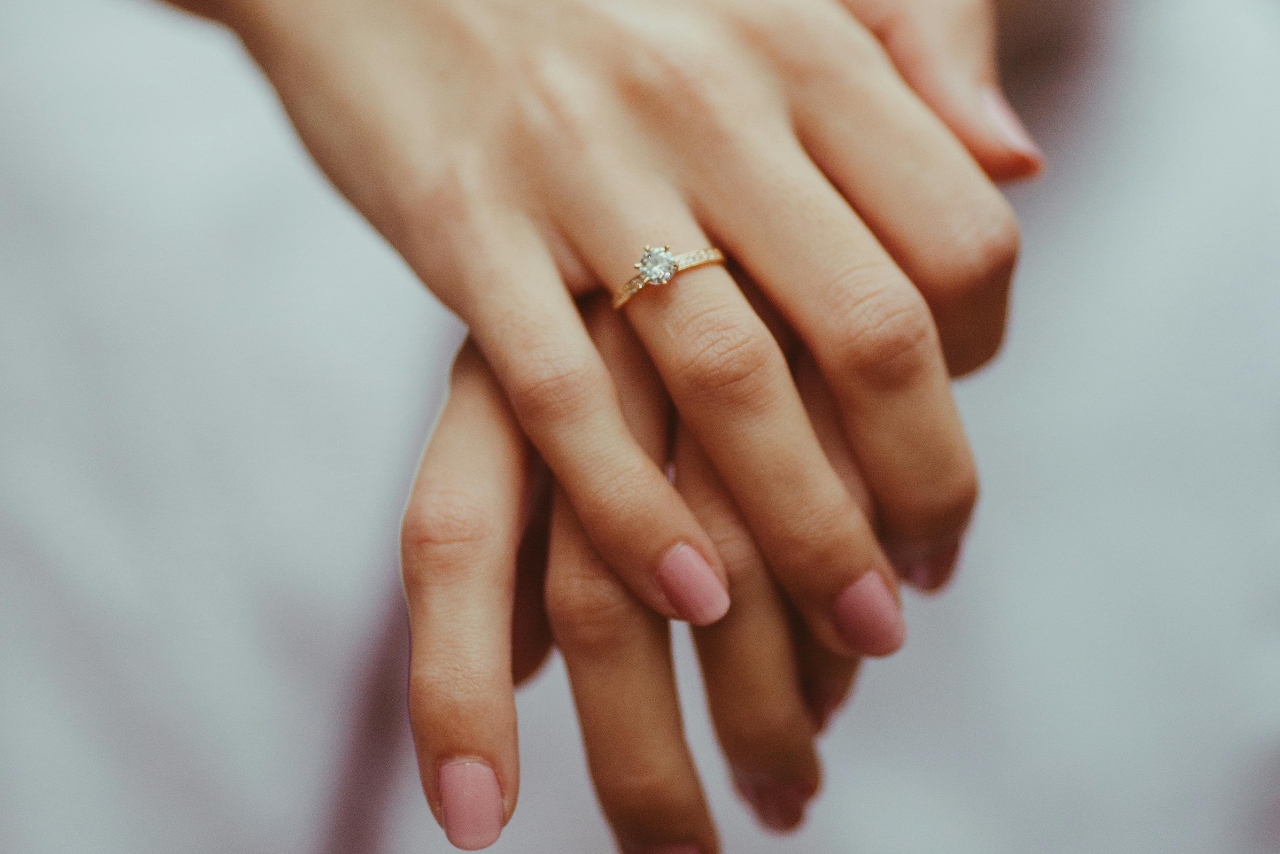 A close up of a yellow gold side stone diamond engagement ring on a woman’s hand with pink manicure.