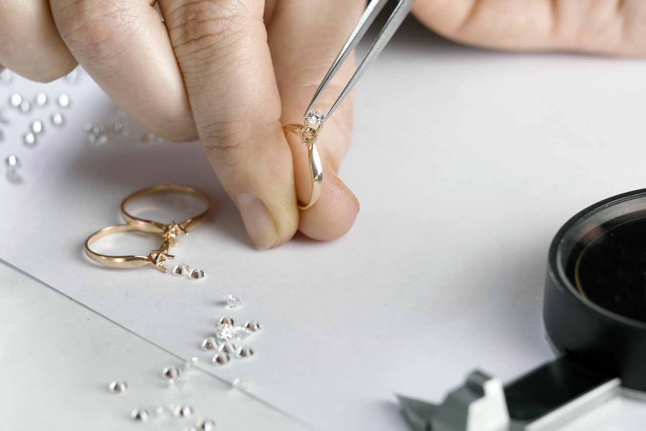 Close-up of a jeweler's hand using tweezers to set a diamond on a gold ring, surrounded by loose diamonds, two rings, and a magnifying glass.