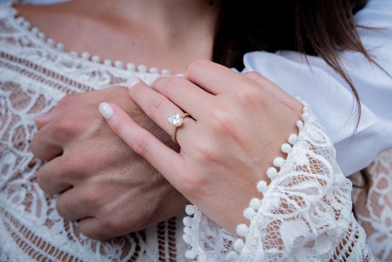 Close-up of a woman in a white lace dress showing a diamond engagement ring on her hand, gently resting on a man's hand.
