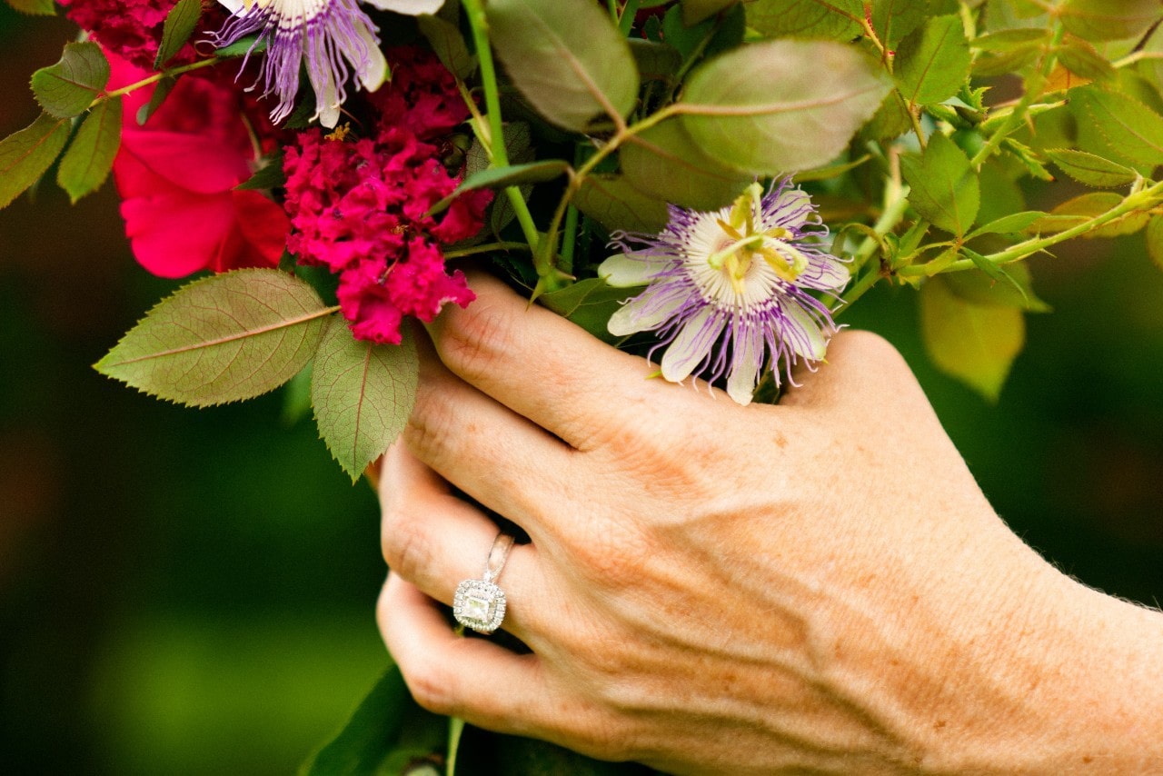 a woman’s hand holding a bouquet of flowers and wearing a halo engagement ring