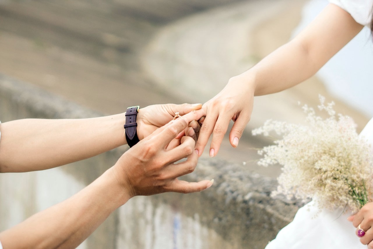 a person sliding an engagement ring on a woman’s finger as she holds a bouquet