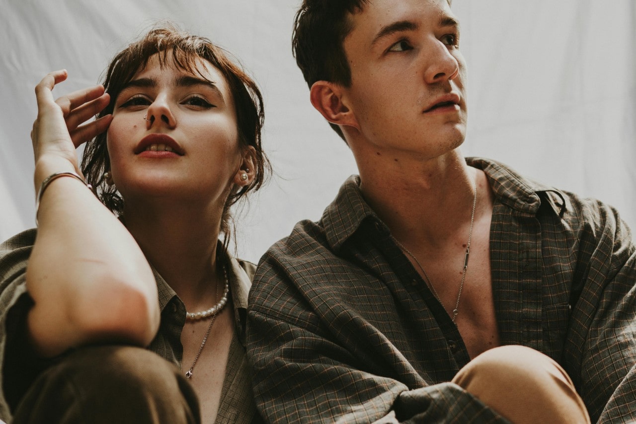 A man and woman posing in button-down shirts, showcasing layered pearl and silver chain necklaces.