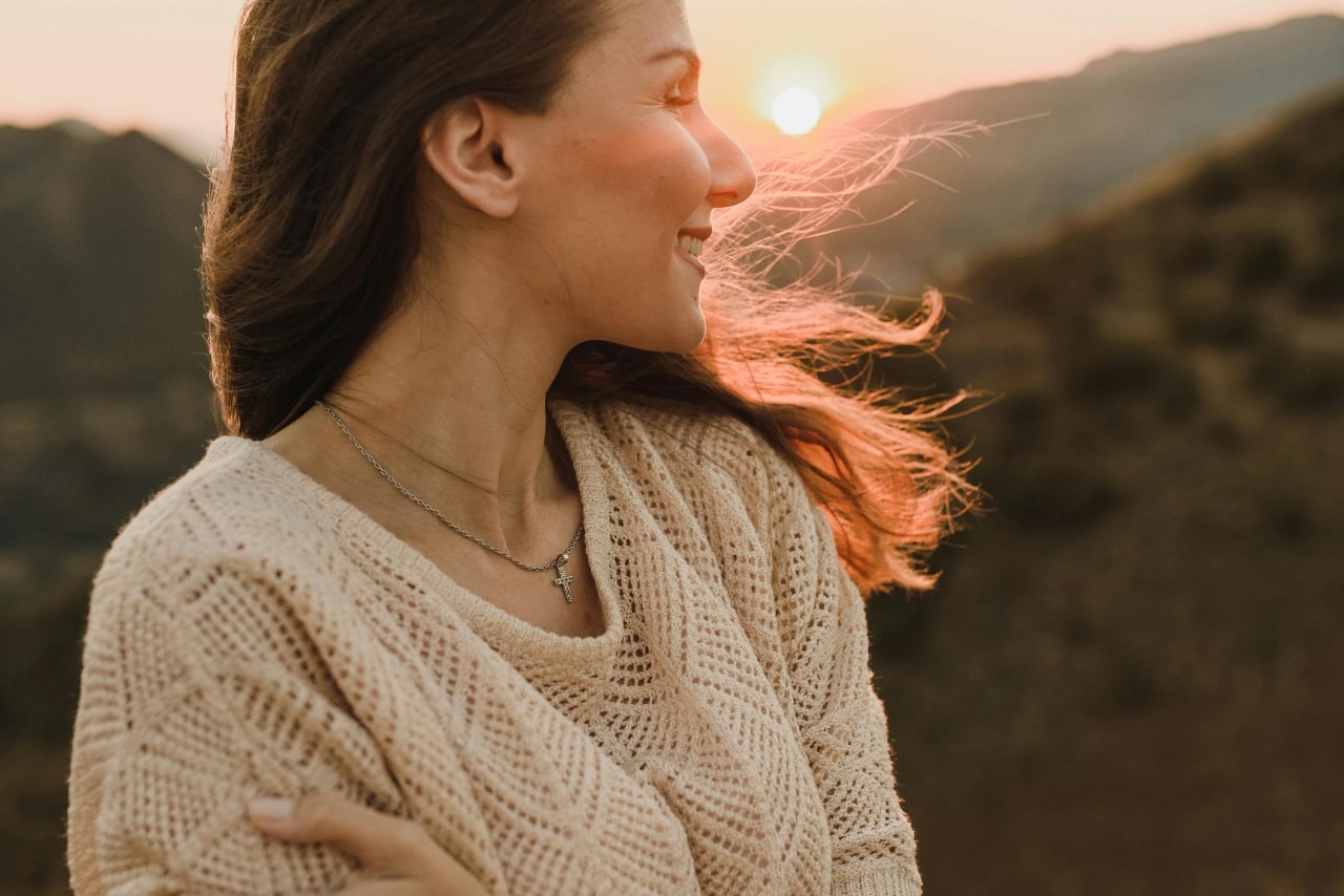 A smiling woman wearing a delicate silver cross necklace during a golden hour mountain sunset.