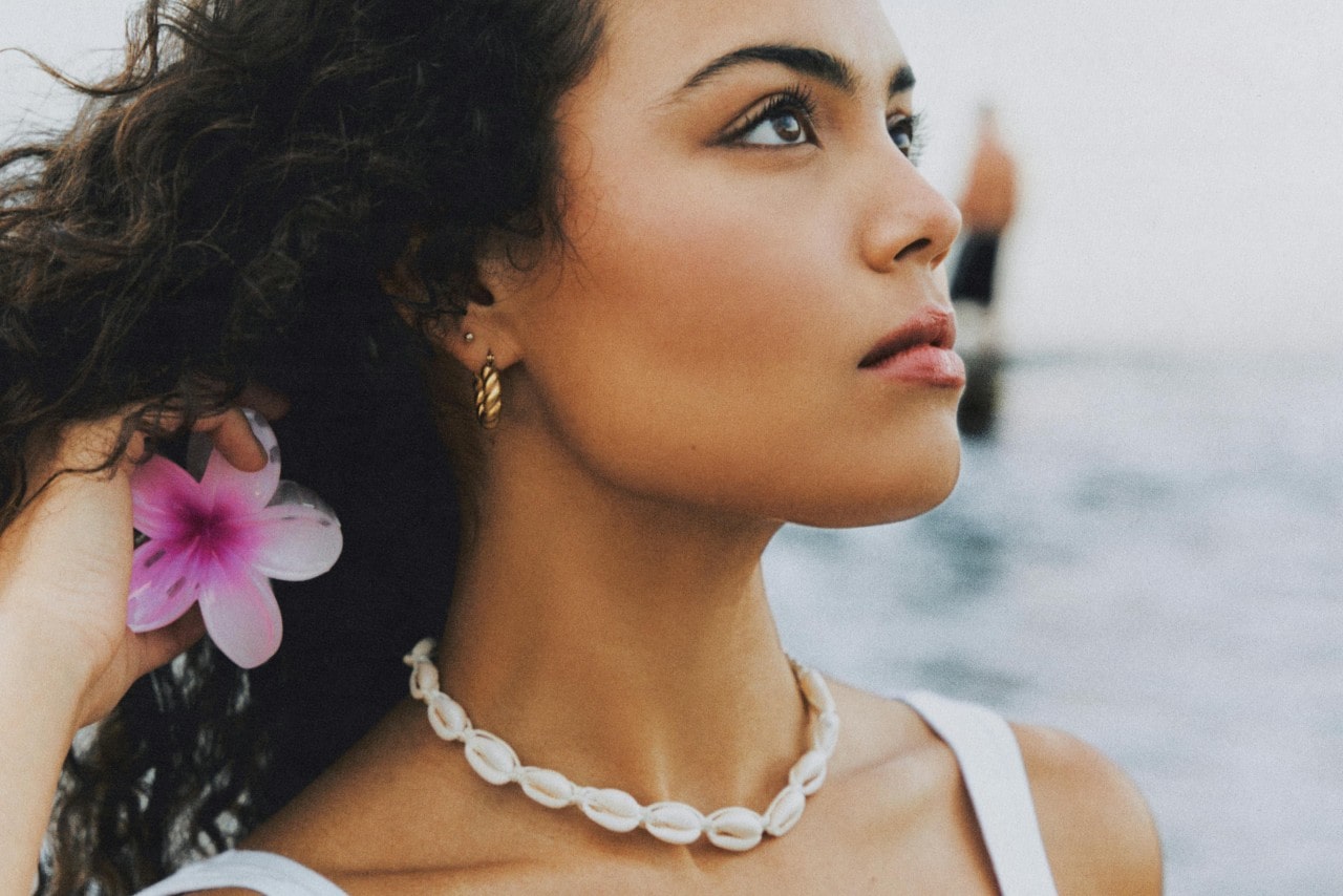 A woman wearing a white cowrie shell choker necklace and gold twisted hoop earrings at the beach.