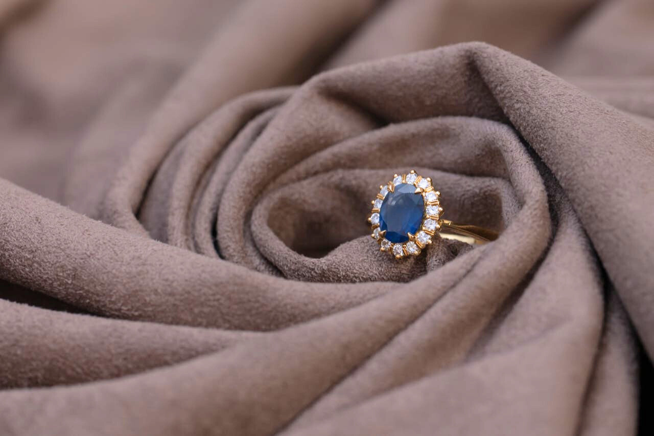A golden engagement ring with a sapphire center stone and a diamond halo, wrapped inside a suede brown fabric.