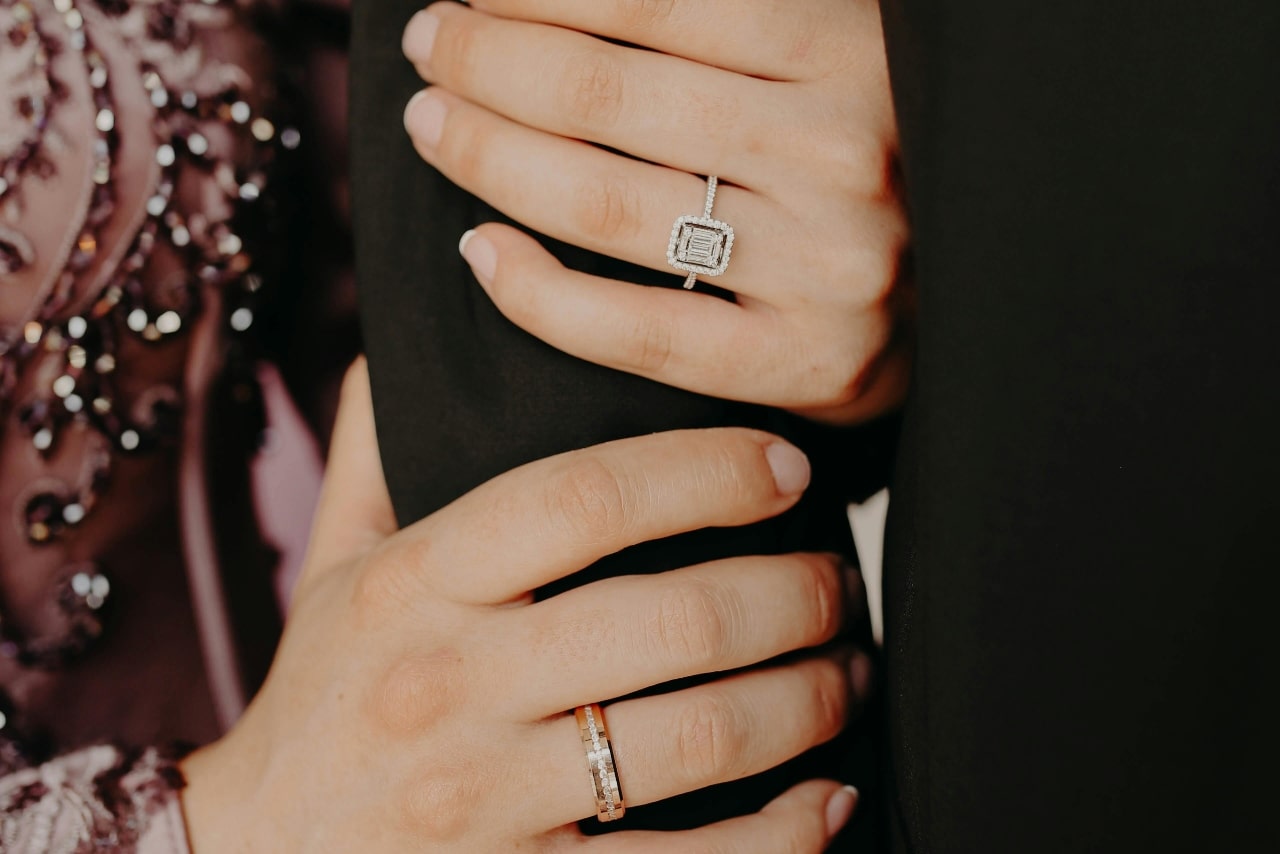 A close up of a woman in a pink sparkling dress holding a men’s hand in a black suit, showcasing an emerald cut diamond halo engagement ring