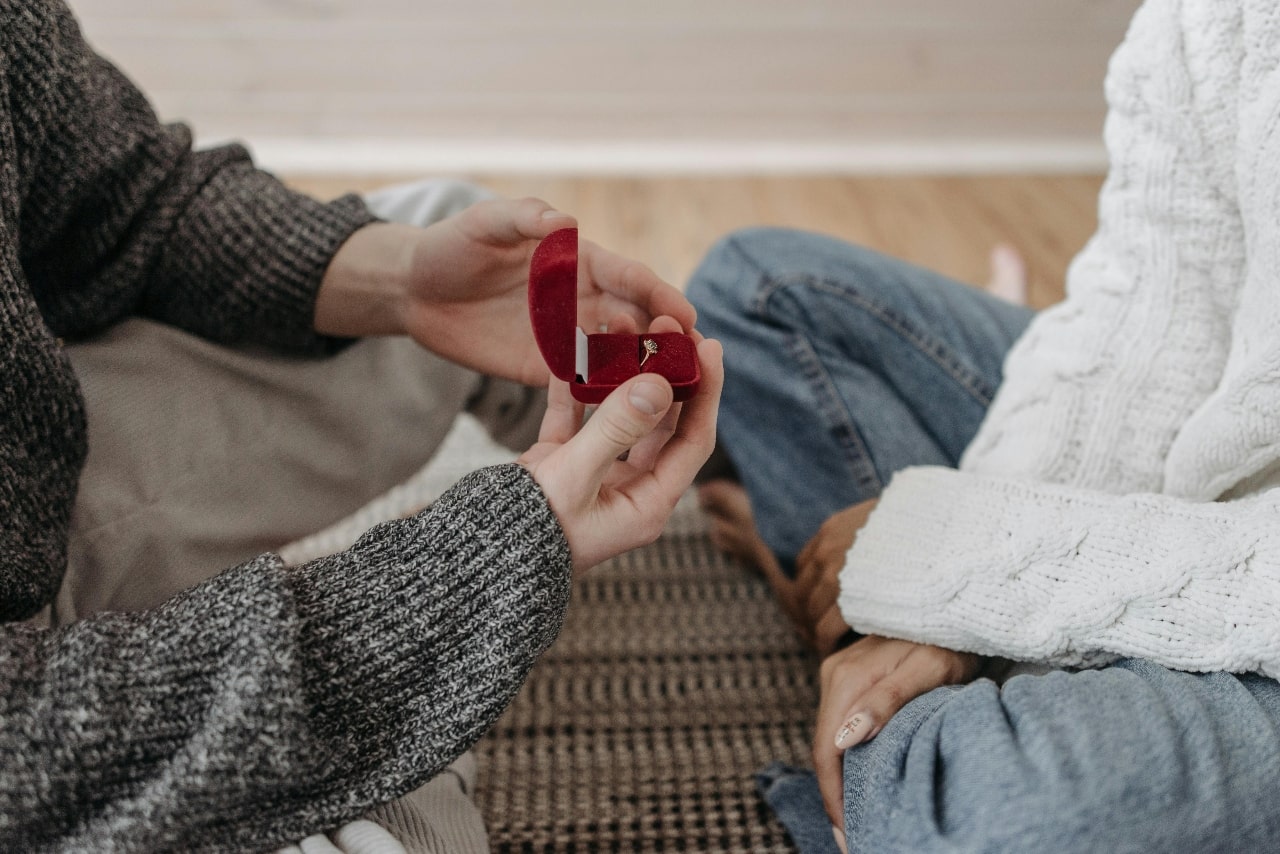 A couple sitting on the floor across from each other, with one holding open a red velvet ring box and proposing to the other.