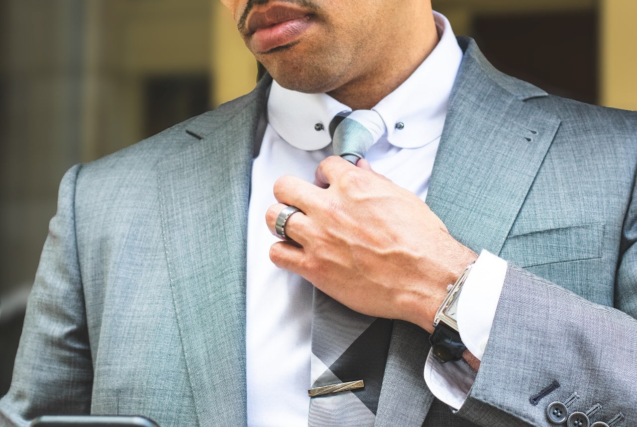 A man in a gray suit adjusts his silver necktie, showcasing a watch and wide wedding band.