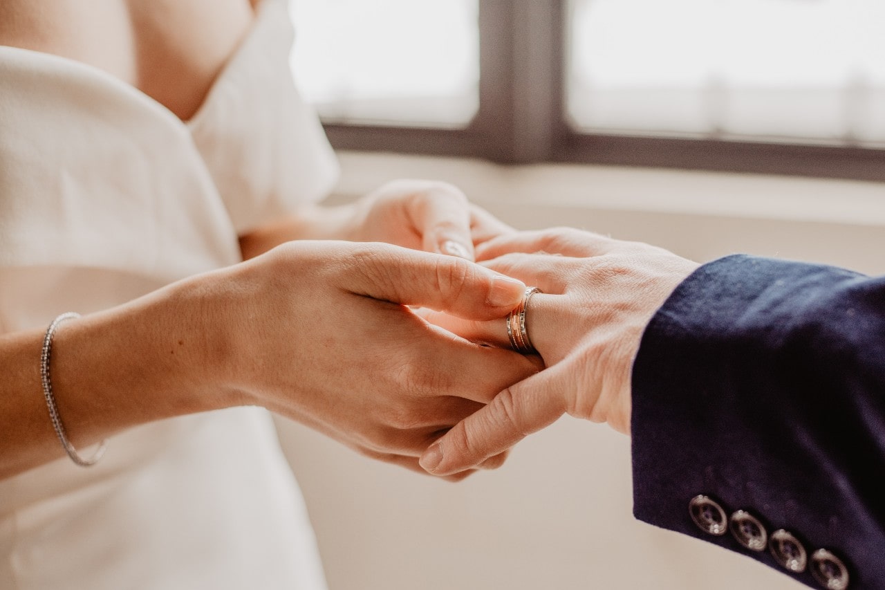 Close-up of a bride in a white dress placing a wedding band on the groom's finger.