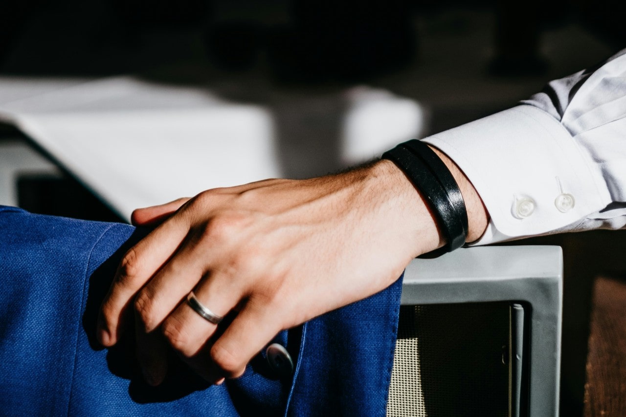 A men’s hand with a silver ring and black bracelet rests on a blue suit jacket draped over a chair.