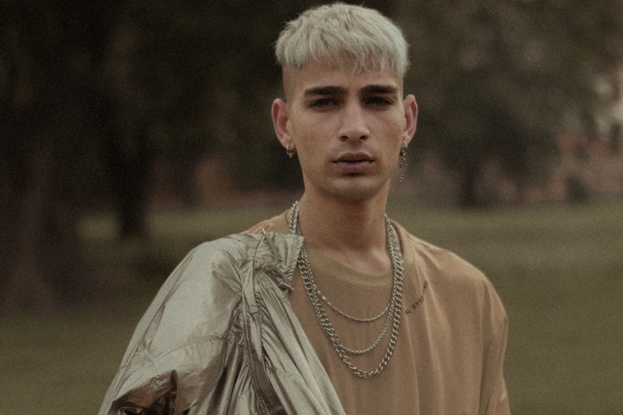 A young man with platinum blonde hair wears layered silver chains, a tan shirt, and a metallic jacket, standing outdoors with a blurred background of trees.