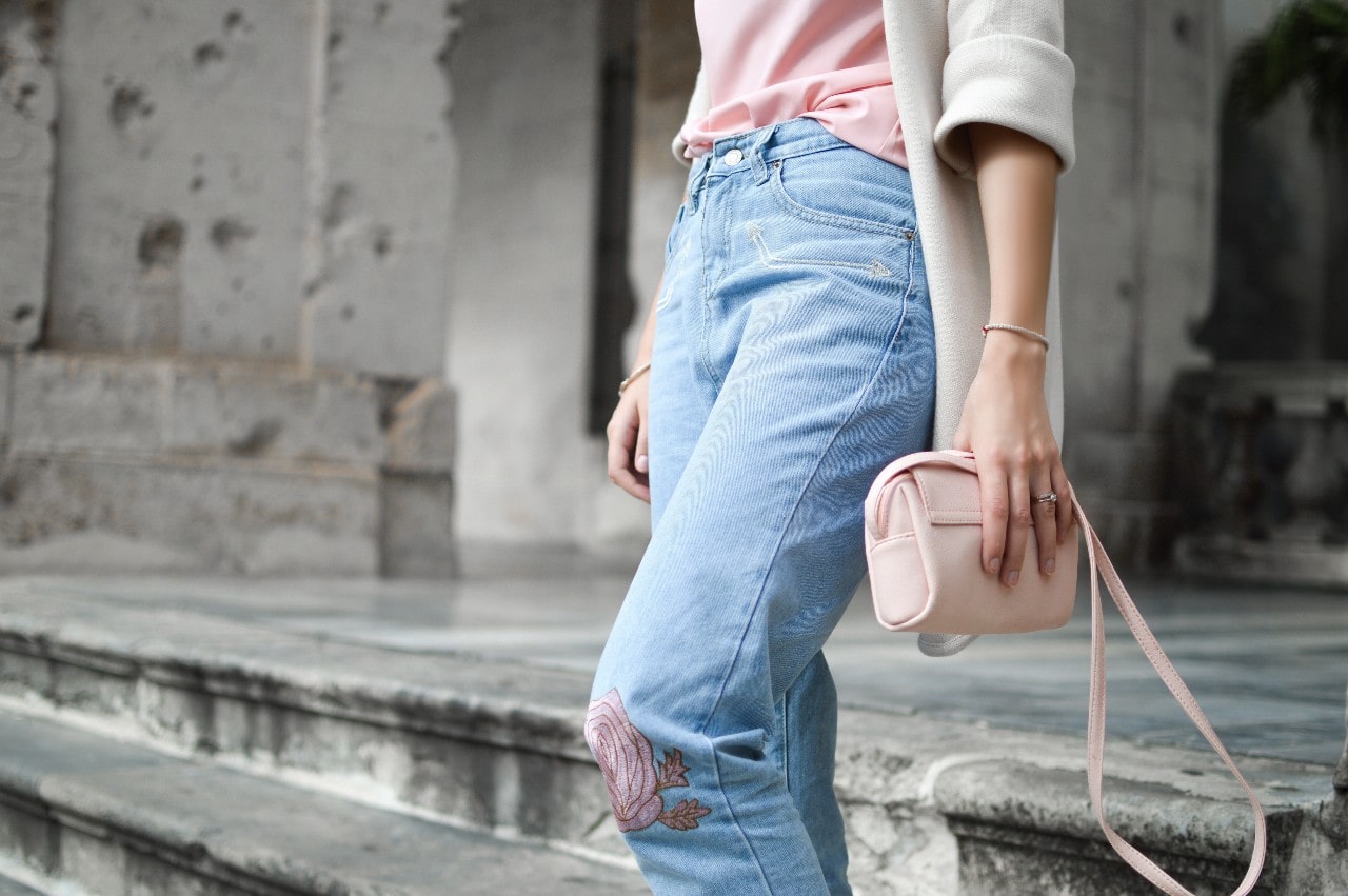 A woman in a pink shirt and blue jeans, holding a pink purse with a small bracelet on her wrist, standing on steps.