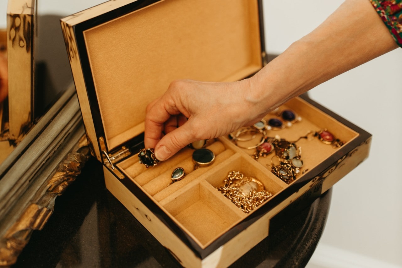 A hand reaching out to a jewelry box that displays an assortment of rings with massive stones, chains, and other fine jewelry.