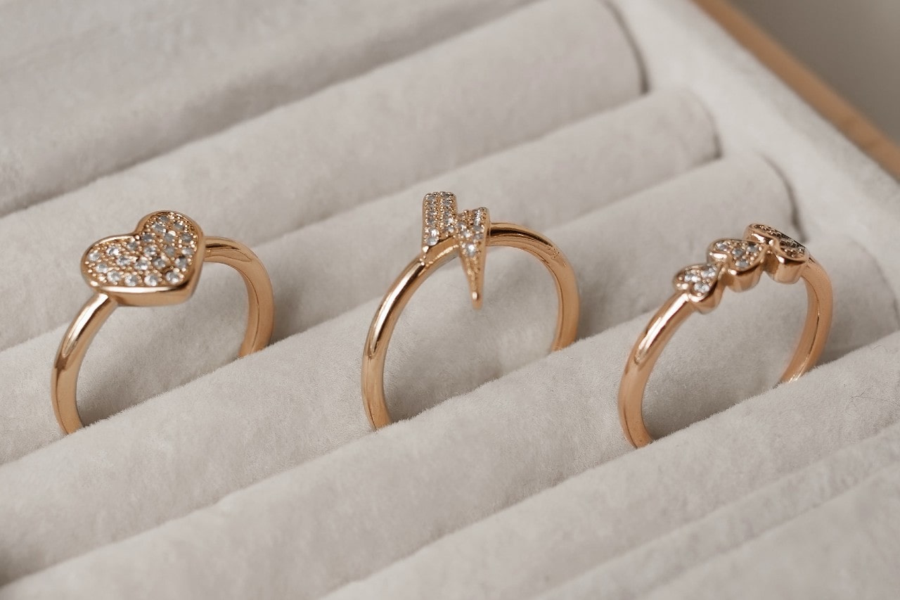 A close-up of 3 yellow gold fashion rings with sparkling diamonds with different center shapes resting in individual slots within a light-colored jewelry display.