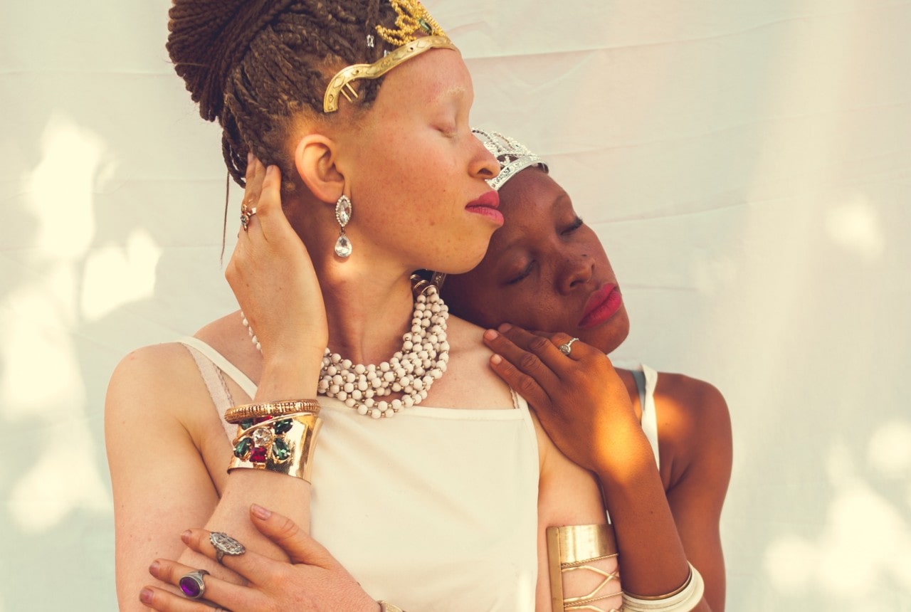 Two women embracing, one laying her head on the other’s shoulder, both adorned with lots of jewelry.