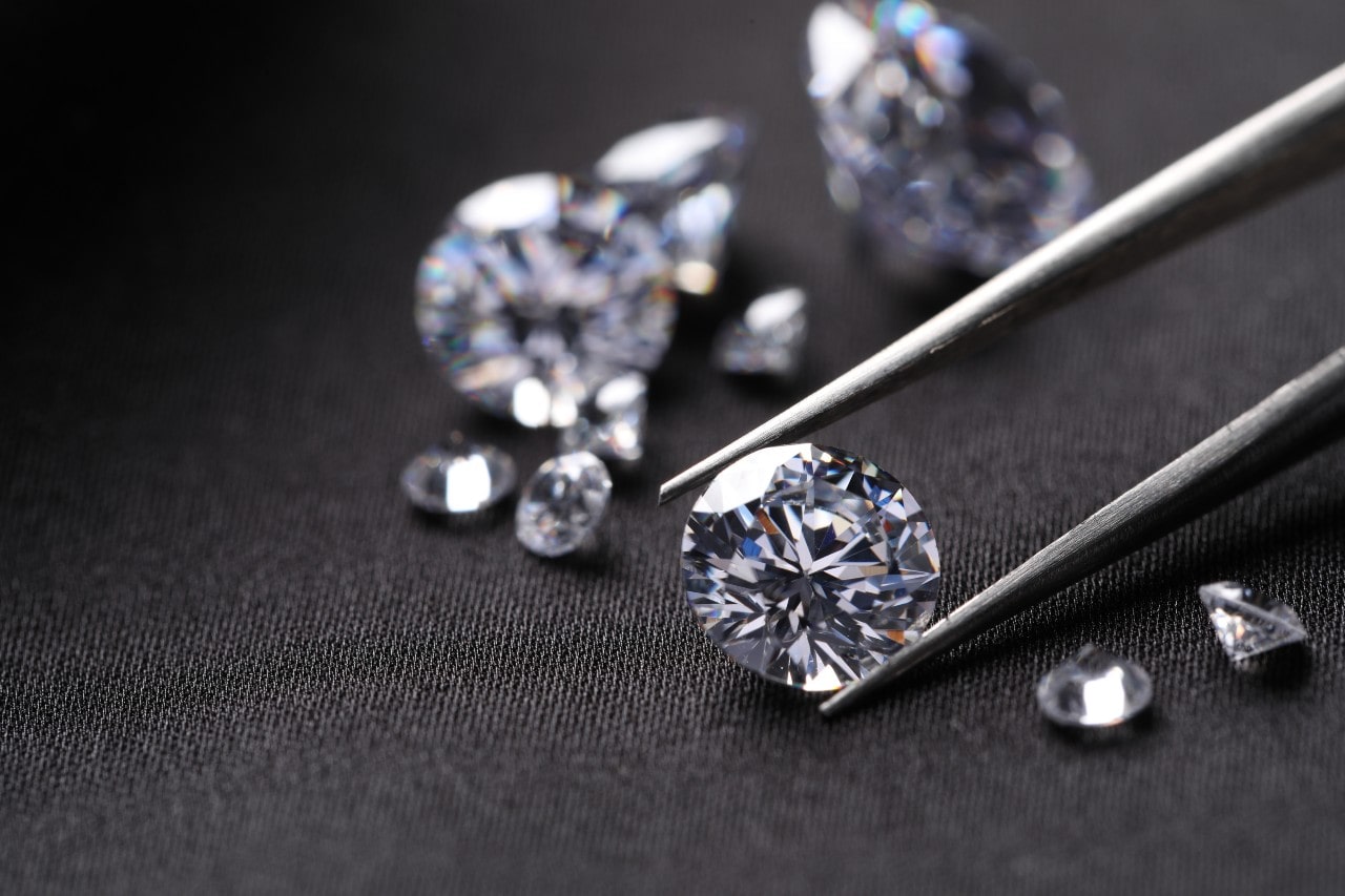 Close-up view of assorted diamonds on dark fabric, with a single stone being inspected using precision tweezers.