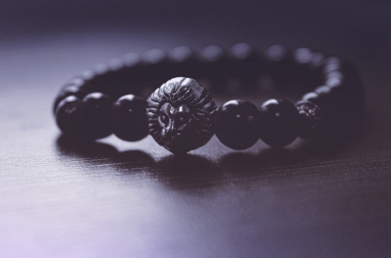 A black beaded bracelet with a detailed lion head centerpiece rests on a dark wooden surface in soft, moody lighting