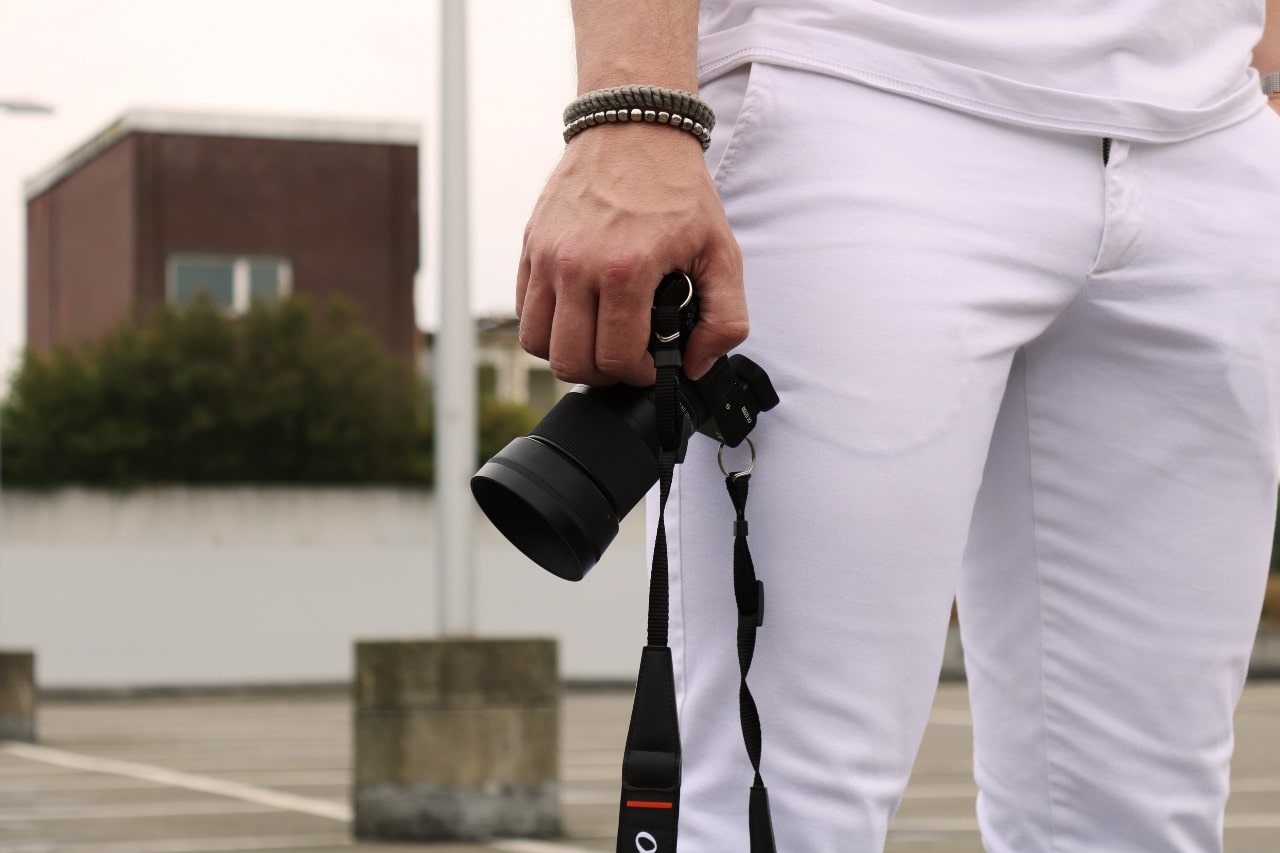 A man in white pants and a white shirt holding a camera at his side, his wrist adorned with stacked beaded bracelets, standing in front of a building.