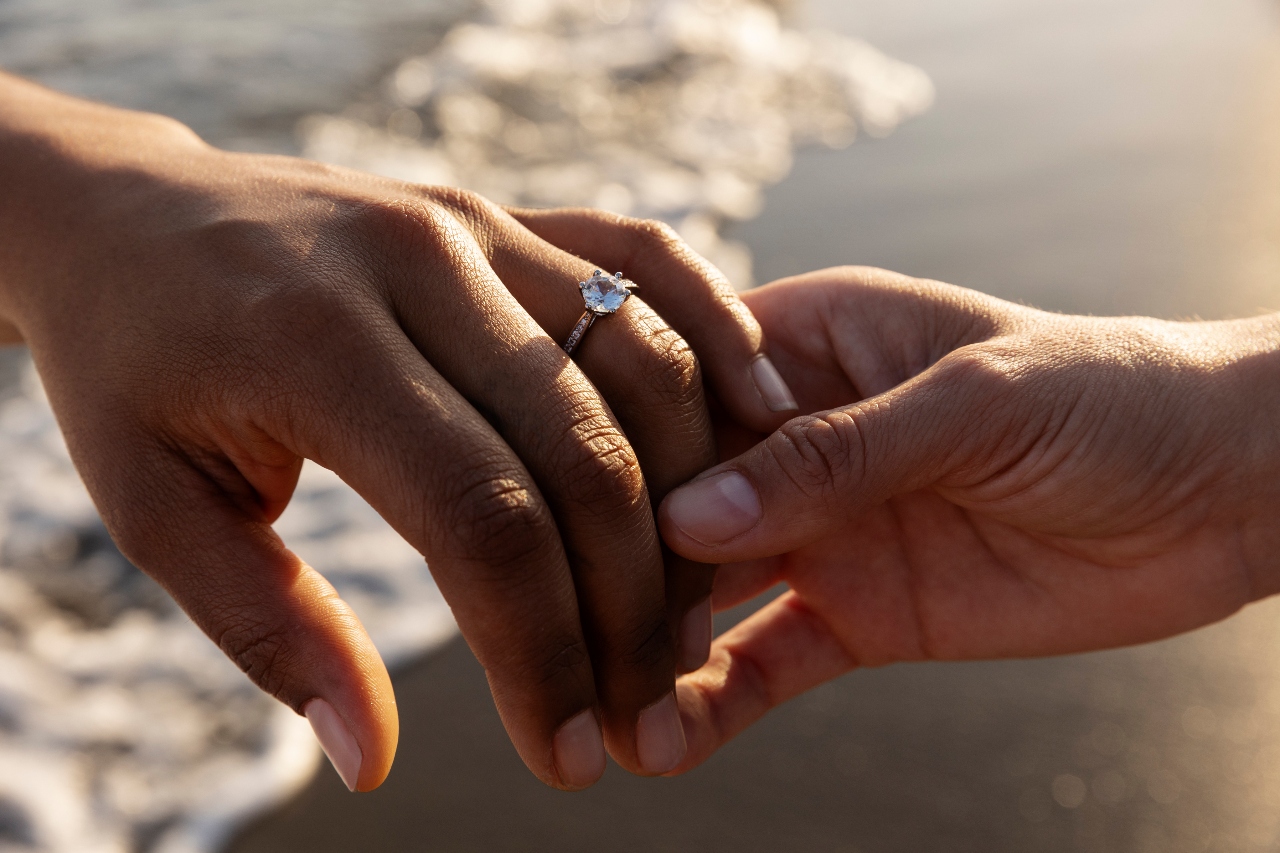 Two hands holding in front of the sea shore, showcasing a diamond engagement ring.