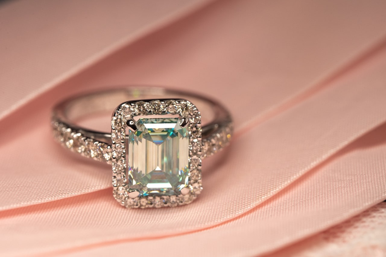 a silver engagement ring featuring a radiant cut center stone and halo setting on a piece of pink fabric a silver engagement ring featuring a radiant cut center stone and halo setting on a piece of pink fabric