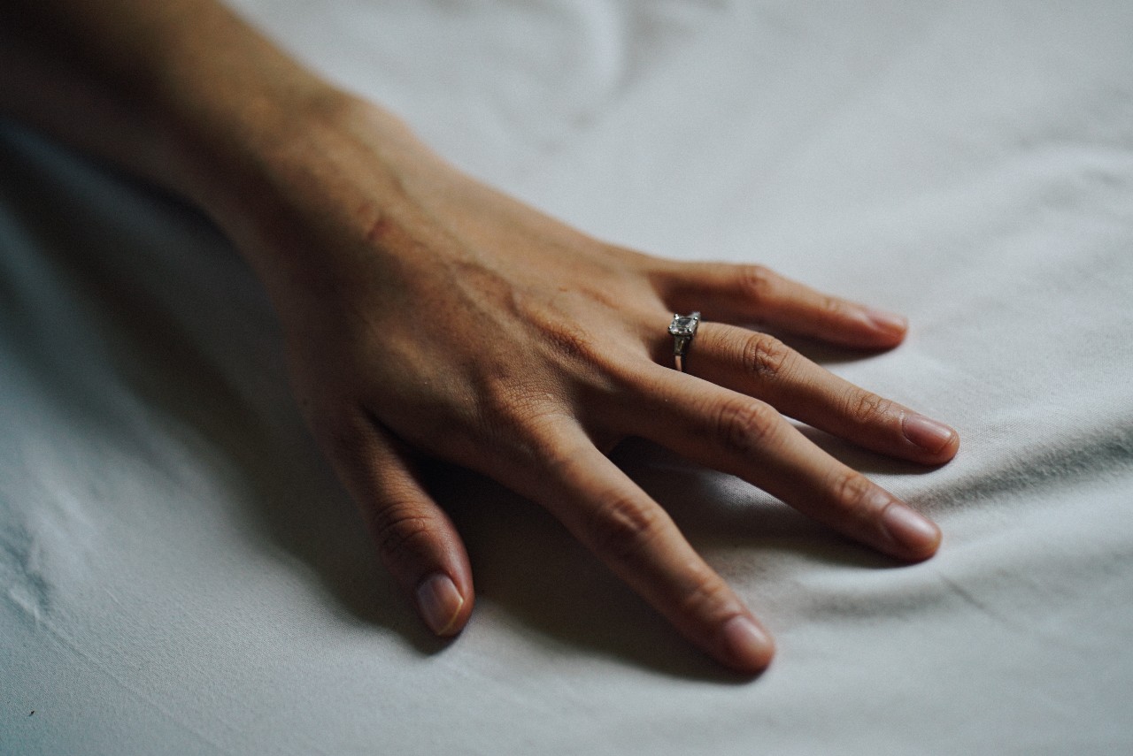 A woman’s outstretched hand with a three stone round cut engagement ring on a white bed sheet.