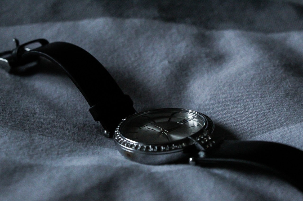 A black watch with roman numerals lays on a gray fabric surface.