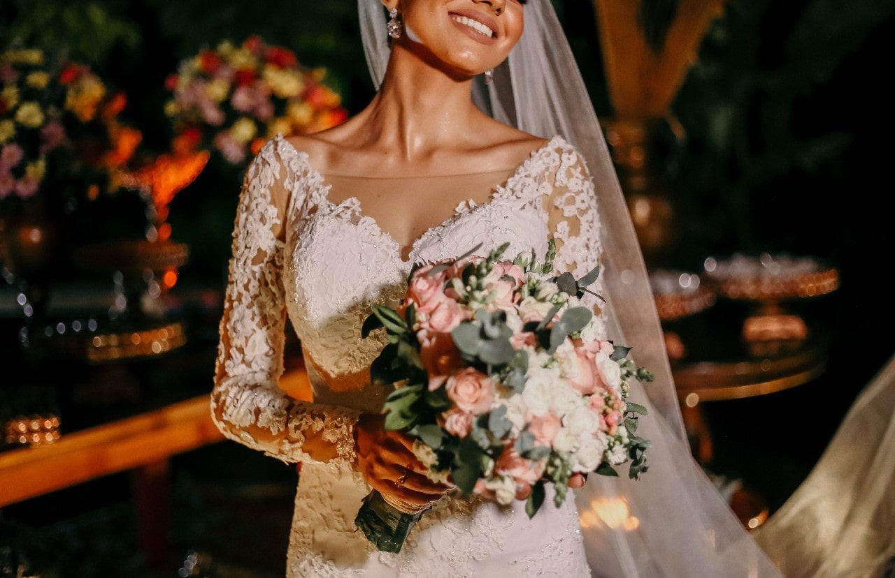 A bride wearing a beautiful wedding band in a lace gown smiles joyfully, holding a pink and white bouquet.