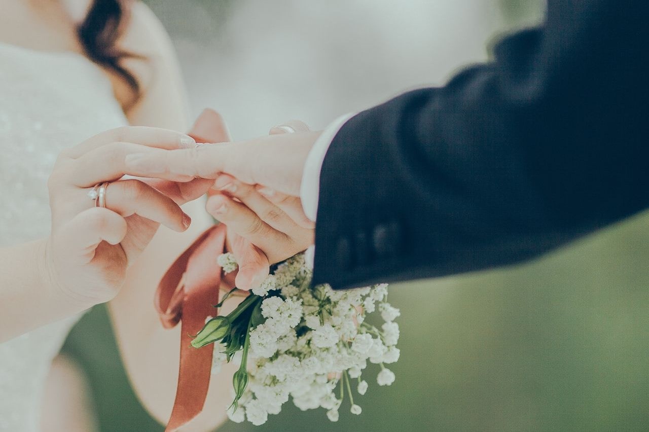 A close-up shot of a bride and groom exchanging wedding rings, the bride wears a white dress and holds a bouquet while the groom is in a black suit.
