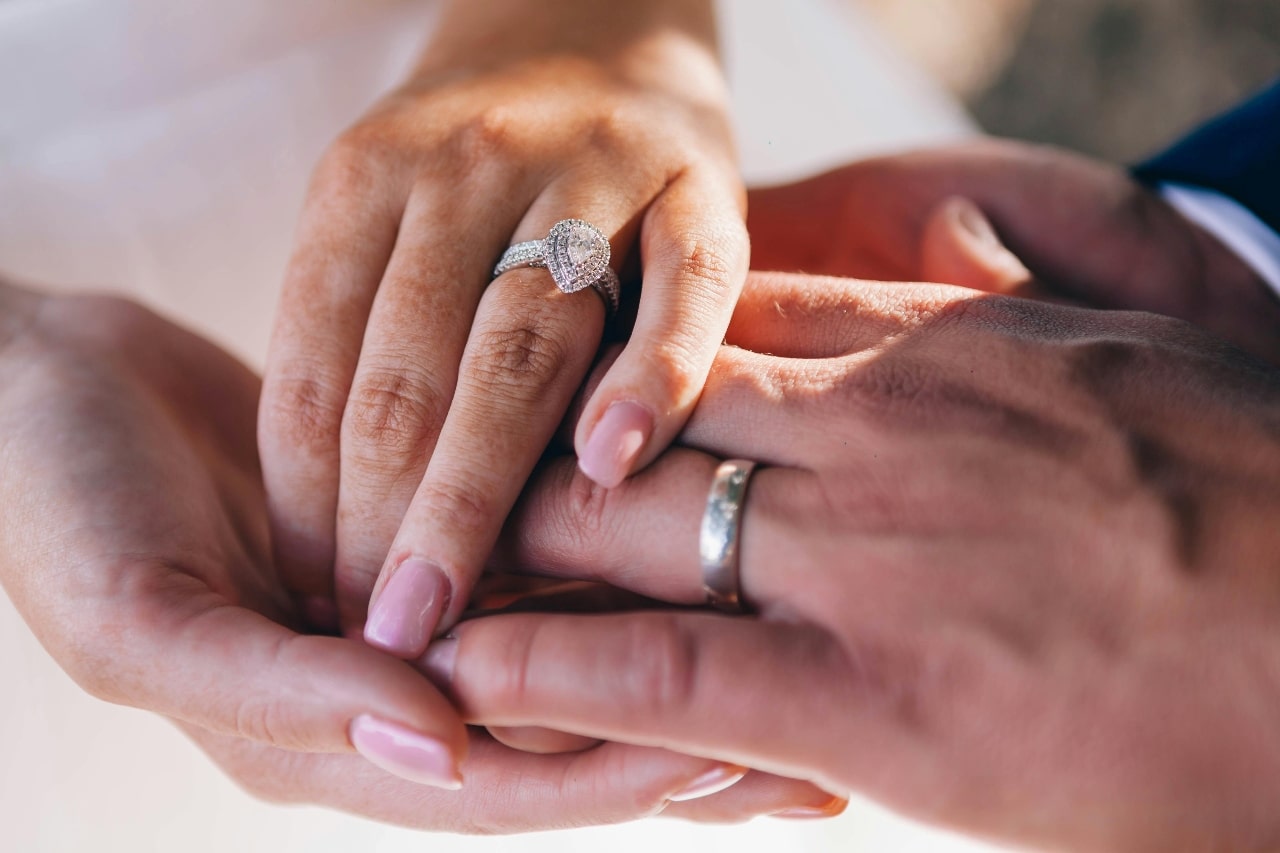 Close-up of a couple holding hands, highlighting a sparkling engagement ring with a halo setting and a simple wedding band.