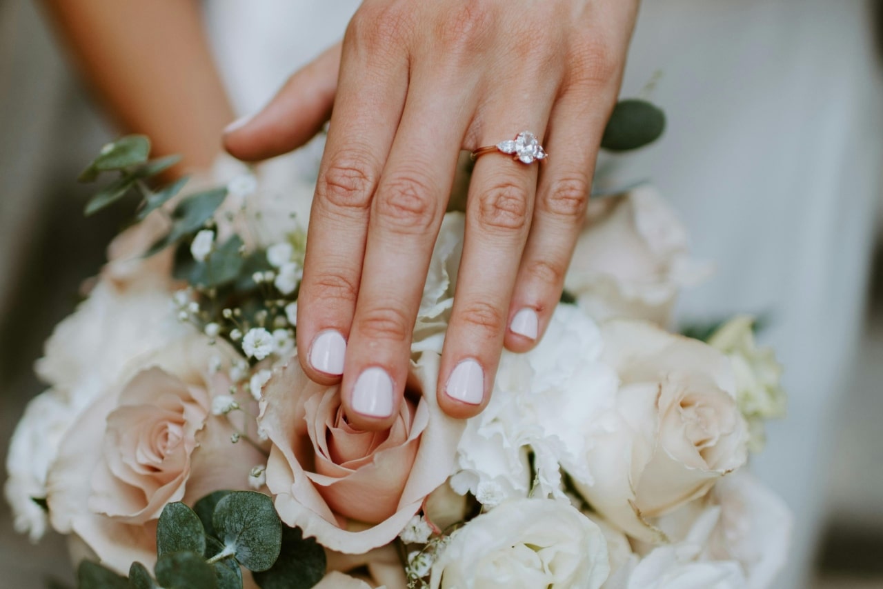 Close up of a hand adorned with a rose gold three stone engagement ring rests on a bouquet of pastel roses and greenery.