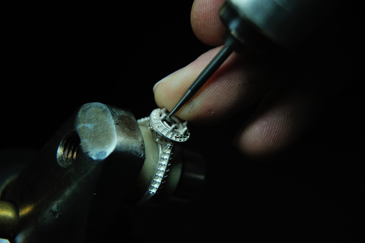 A close-up of a craftsman using a tool to secure a diamond setting in an intricate white gold engagement ring
