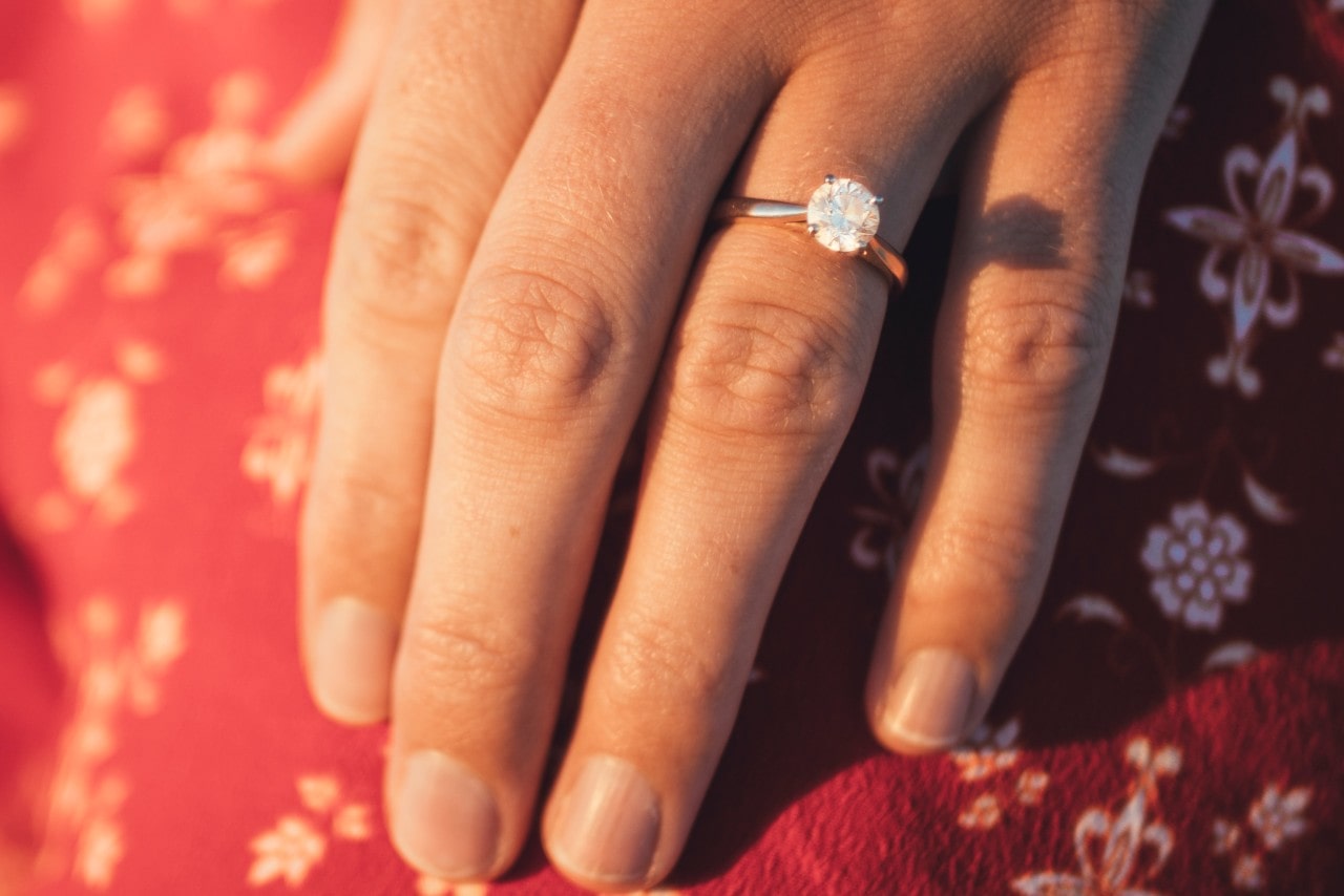 a woman’s hand resting on red fabric, adorned with a diamond engagement ring