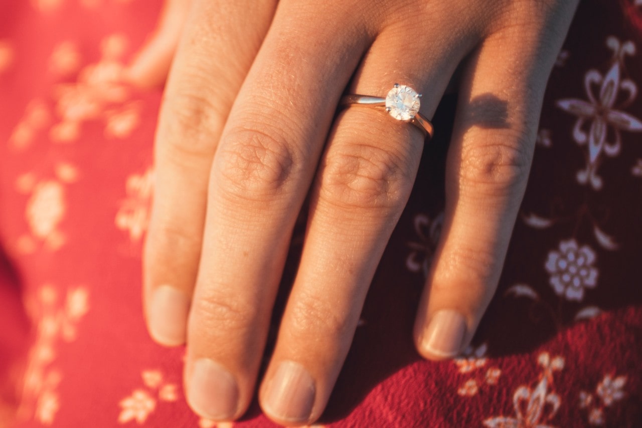 a woman’s hand resting on red fabric, adorned with a diamond engagement ring a woman’s hand resting on red fabric, adorned with a diamond engagement ring
