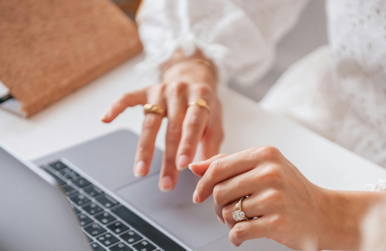 A close up of a woman’s hands typing on a laptop, with emphasis on her yellow gold oval cut engagement ring, along with textured fashion rings on her right hand. A close up of a woman’s hands typing on a laptop, with emphasis on her yellow gold oval cut engagement ring, along with textured fashion rings on her right hand.
