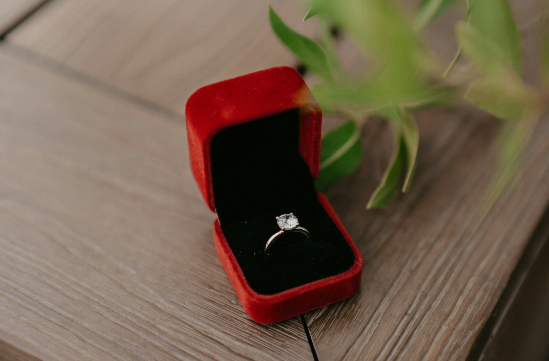 A classic round-cut diamond engagement ring displayed in a red velvet box atop a wooden surface. A classic round-cut diamond engagement ring displayed in a red velvet box atop a wooden surface.