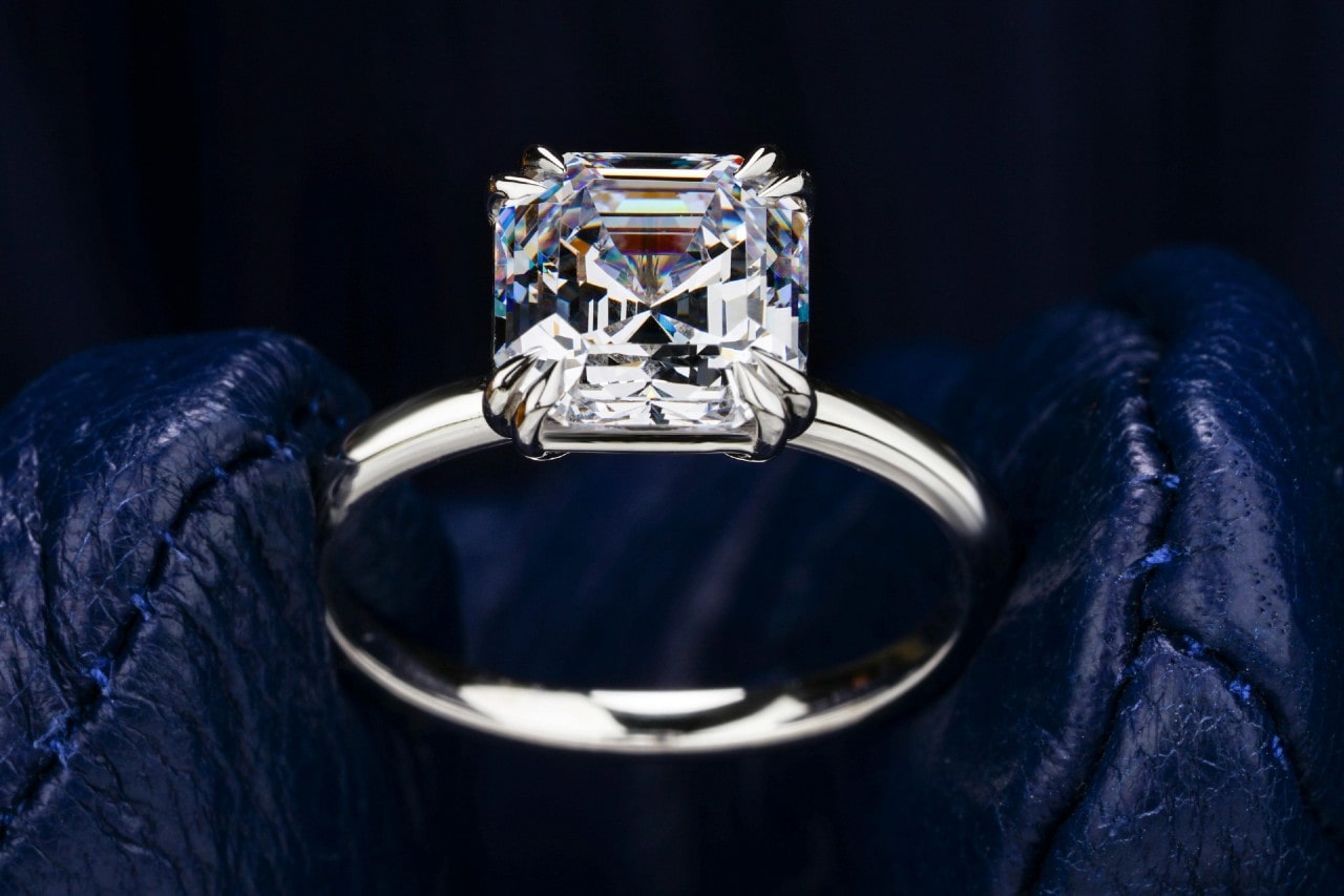 An cushion-cut diamond solitaire engagement ring set in a sleek white gold band is displayed against deep blue leather gloves. An cushion-cut diamond solitaire engagement ring set in a sleek white gold band is displayed against deep blue leather gloves.